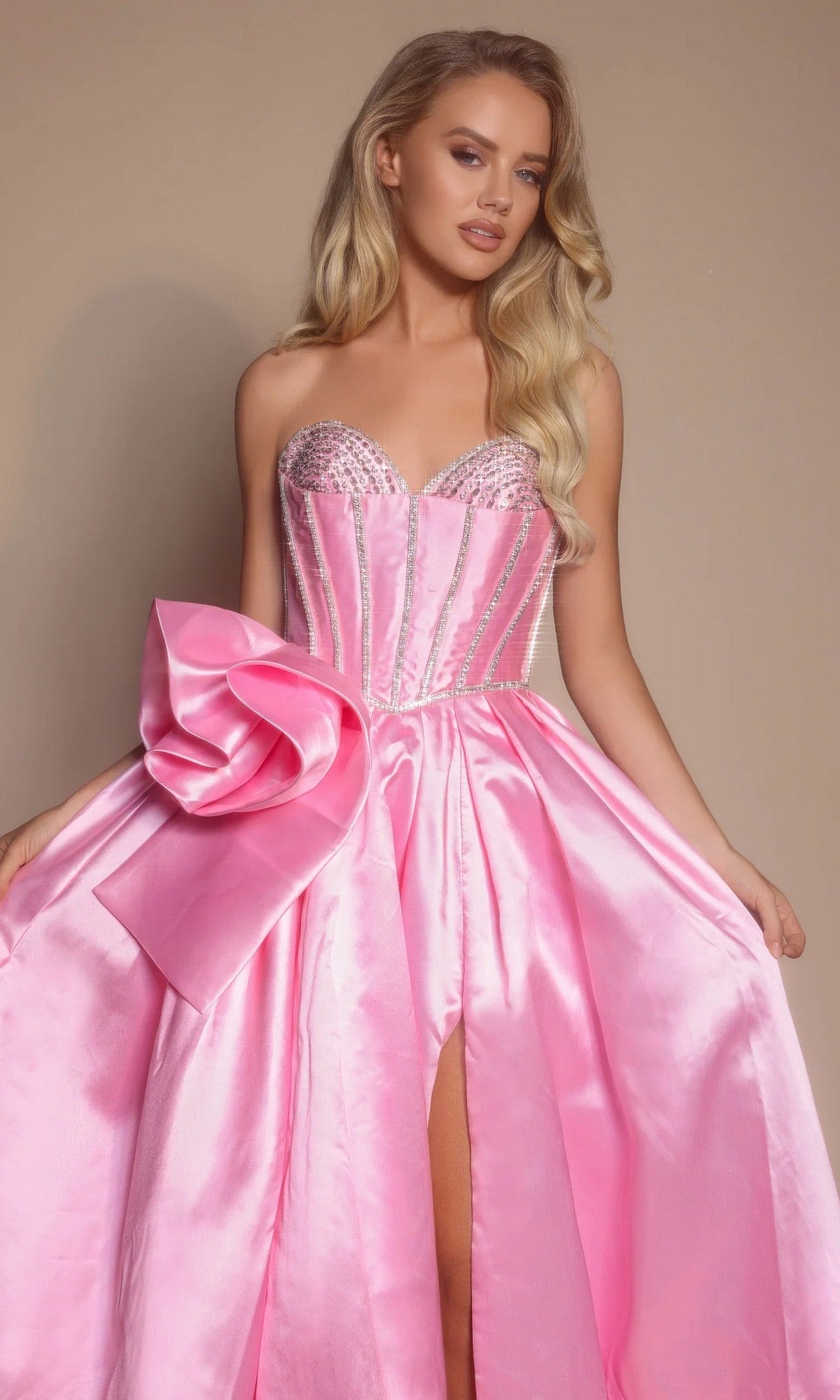 A woman wears the Portia and Scarlett PS26079, a strapless satin long formal dress with a large bow, beaded corset bodice, and thigh-high slit, standing against a neutral background.